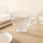Przezroczysta szklanka z pionowymi żłobieniami na drewnianym blacie, obok druga szklanka i talerz, idealne do kawy lub herbaty, elegancki i nowoczesny design.