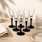 Przezroczyste kieliszki do szampana z czarną podstawą, eleganckie szkło na uroczystości, na stole z białą tkaniną i ceramicznymi naczyniami, nowoczesny design.