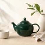 Przedmiotowa czarna czajnik ceramiczny na drewnianym stole z białą ceramiką i zielonymi liśćmi w tle, elegancki i nowoczesny design, idealny do kuchni i serwowania napojów.